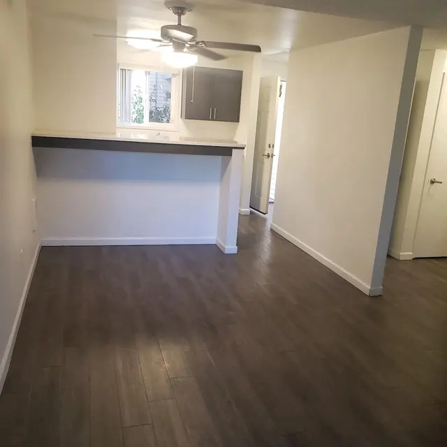 A modern open-concept living space featuring wood-like flooring, a ceiling fan, and a bar counter with an adjacent window letting in natural light.