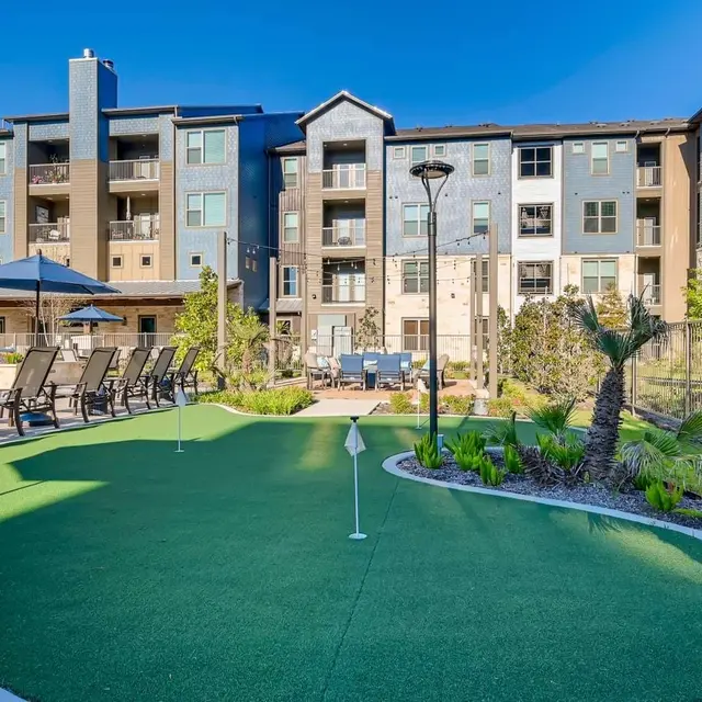 A well-maintained apartment complex featuring a green putting green area with lounge chairs and a view of the buildings in the background.