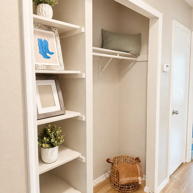 A small closet space featuring white shelves with decorative items, a green cushion on a shelf, and a wicker basket on the floor.