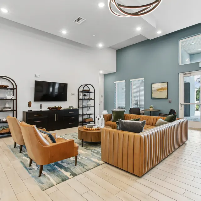Modern lounge area featuring a brown leather sofa, armchairs, a coffee table, and a wall-mounted TV with large windows.