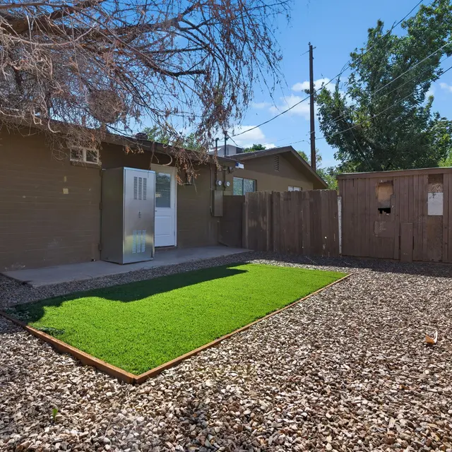 A backyard featuring a patch of artificial grass surrounded by gravel, a wooden fence, and a house exterior in the background.
