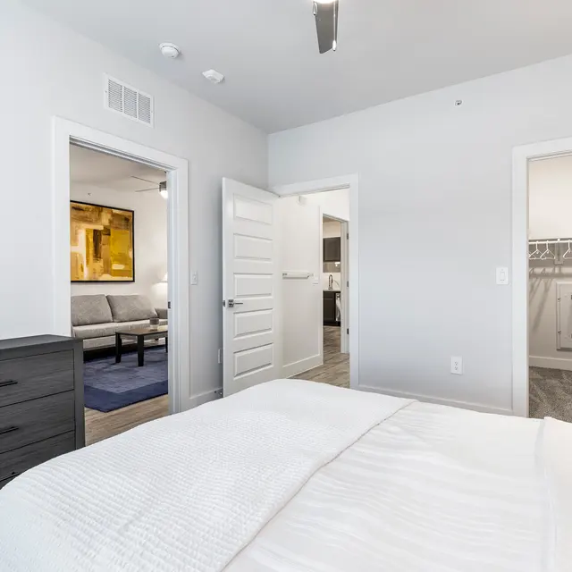 A modern bedroom featuring a bed with white bedding and a black dresser. Open doors reveal views into adjoining rooms, including a living area with a sofa and a dining space. The walls are painted in light colors with minimalistic decor.