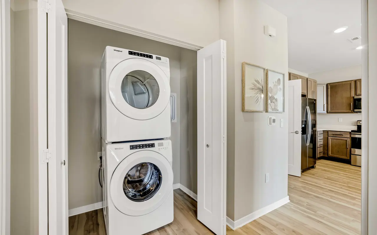 A modern laundry room featuring a stacked washer and dryer unit, with light-colored walls and wooden flooring. Two white doors lead to the laundry area, and there are framed artworks on the wall near the entrance.
