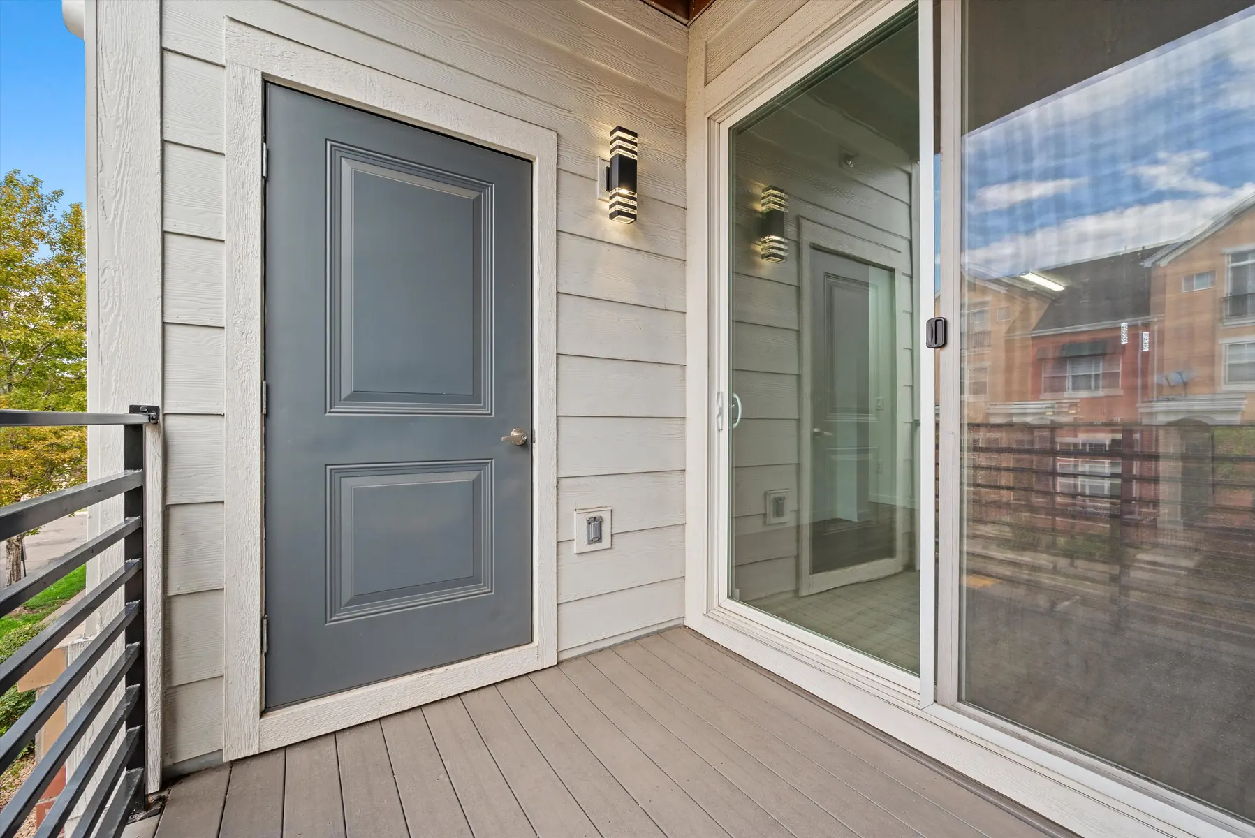 A balcony area featuring a gray door and sliding glass door, overlooking a landscaped area with trees.