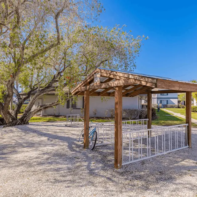 Outdoor Pavilion in a Park Setting A wooden pavilion with a gravel floor surrounded by green lawns and a large tree, with buildings in the background under a clear blue sky.