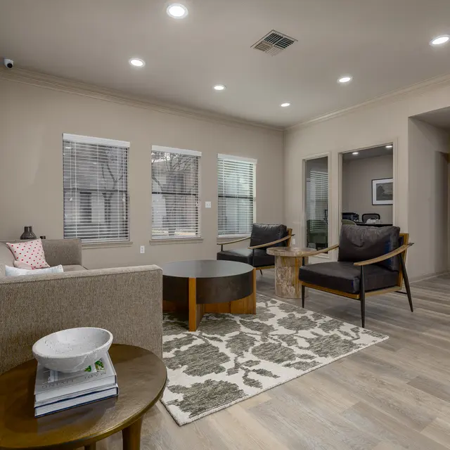 A modern living room featuring a light-colored sofa, two dark wooden chairs, a round coffee table, and a patterned area rug. The room has multiple windows allowing natural light to fill the space, with a light-colored wall and wooden flooring.