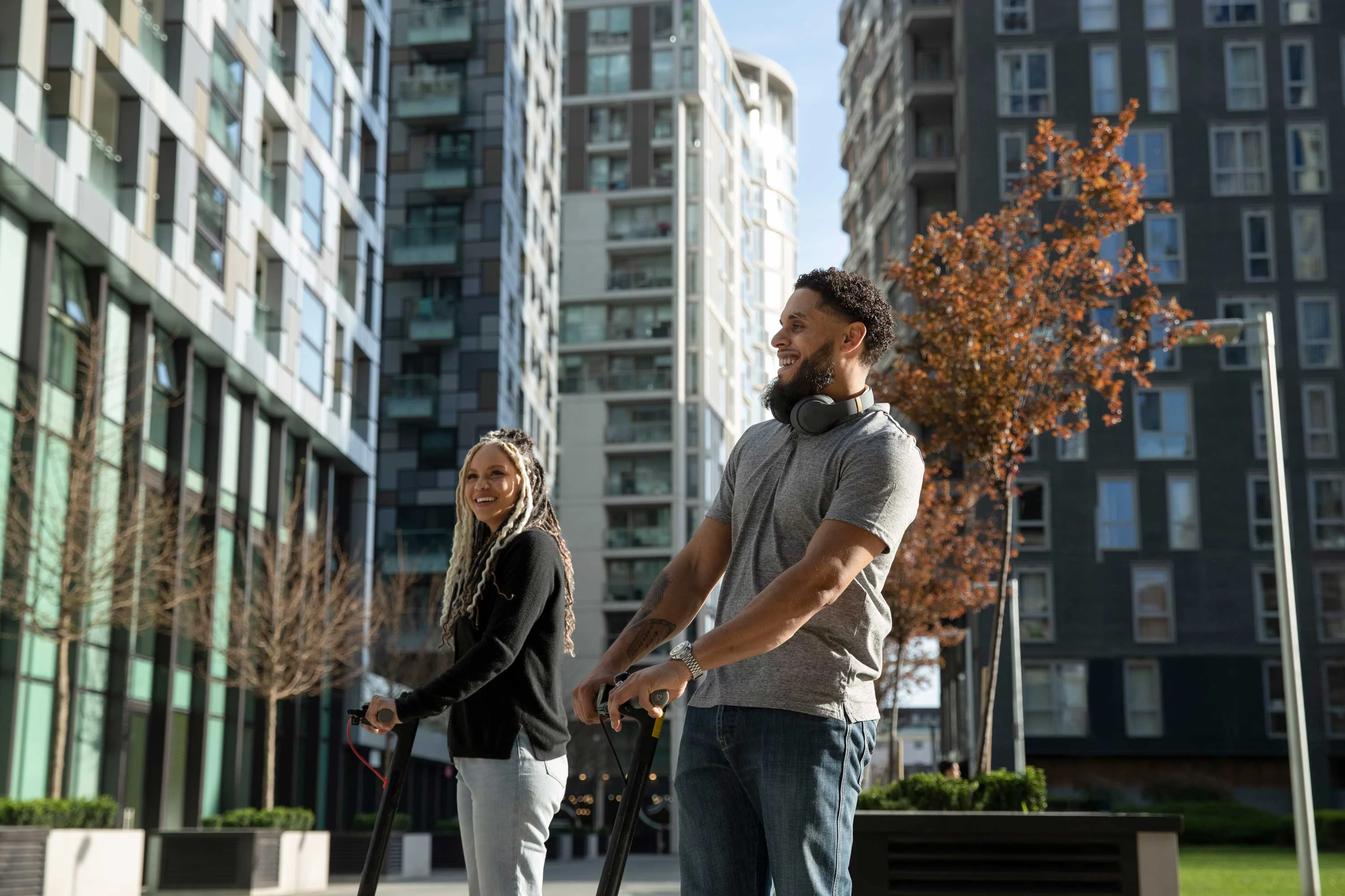 A man and a woman are standing outside modern apartment buildings, both holding skateboards and smiling. The woman has long braided hair, and the man is wearing headphones. The surroundings include trees and contemporary architecture.