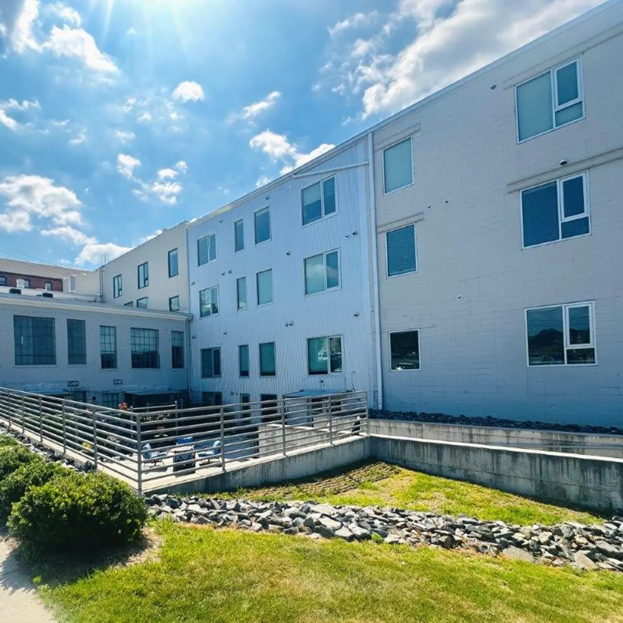 Exterior view of a modern white building, featuring multiple windows, surrounded by greenery and a walkway, under a bright blue sky with clouds.