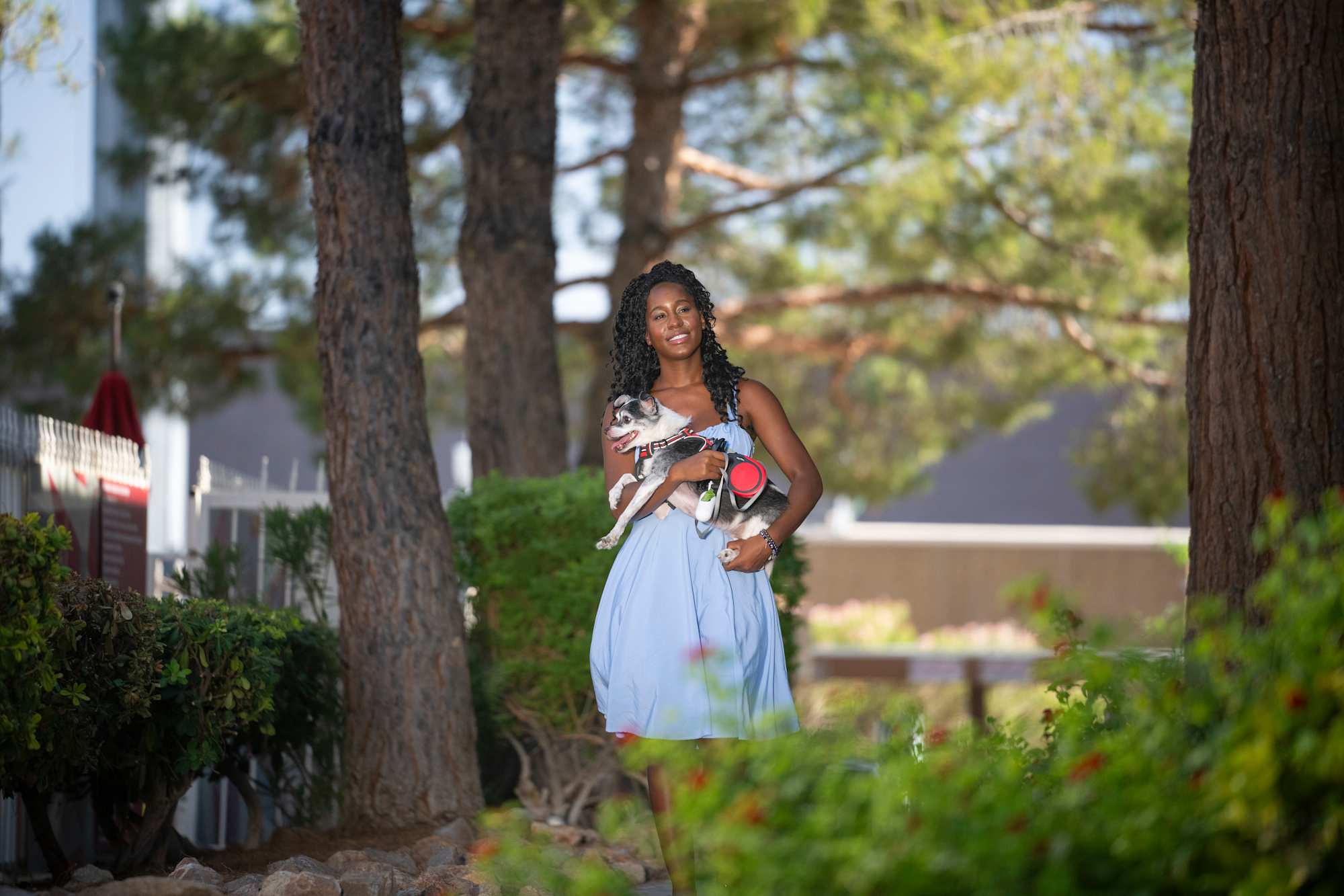 Woman Enjoying a Day Outdoors with Her Dog A woman in a light blue dress stands outdoors, holding a small dog in her arms amidst trees and greenery.
