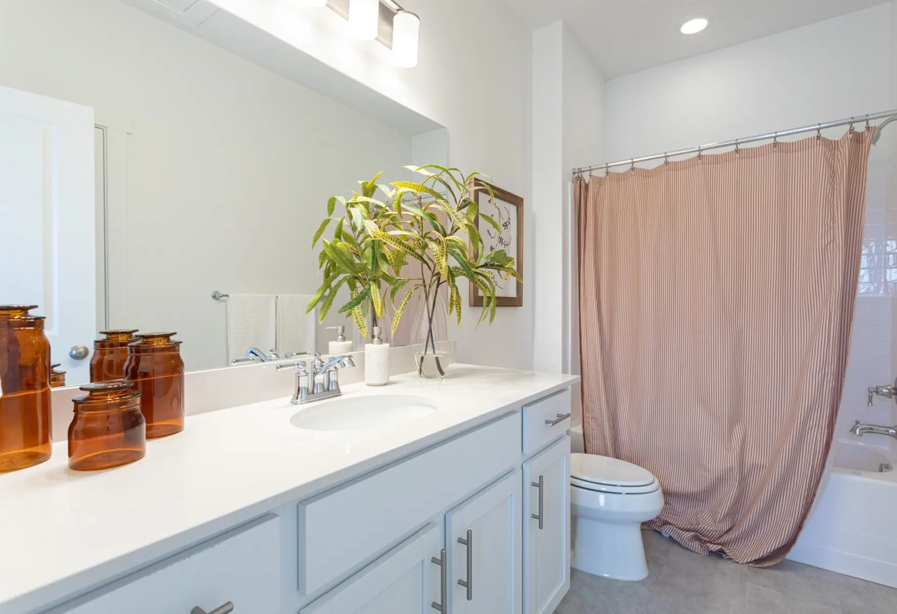 Modern Bathroom Interior A modern bathroom featuring a white countertop with amber jars, a potted plant, and a large mirror above. There is a shower area with a light pink curtain and a white toilet to the side.