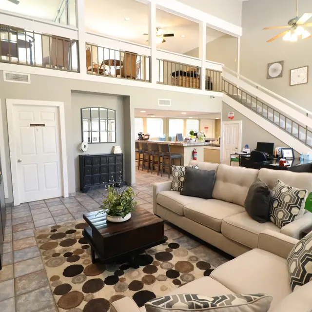 Spacious Modern Living Room A spacious living room featuring a large, light-colored sectional sofa with decorative pillows, a coffee table on a patterned rug, and a view of a loft area. The room has high ceilings with a ceiling fan, natural light coming in from large windows, and a staircase leading to the upper level. There is also a desk area and a bar counter visible in the background.