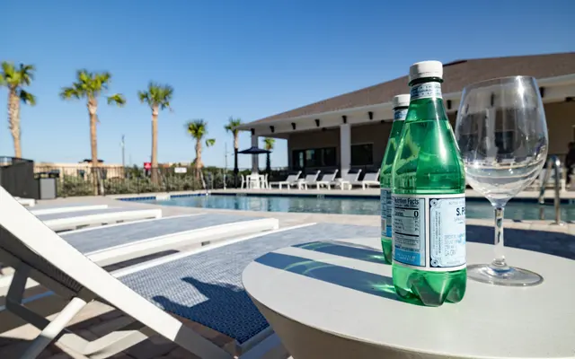 A poolside scene featuring a glass of sparkling water and a wine glass on a small table, with lounge chairs and palm trees in the background.