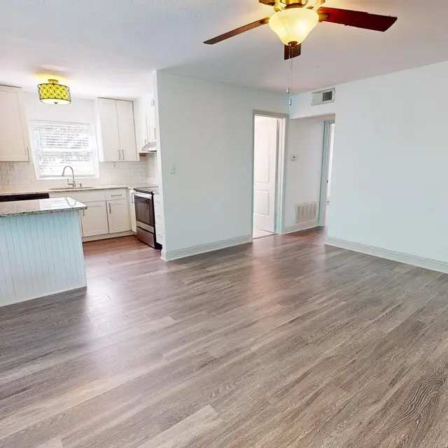 A spacious, modern kitchen and living area with wooden flooring. The kitchen features stainless steel appliances and light-colored cabinetry, with a window providing natural light. A ceiling fan is present, and the area is well-lit.