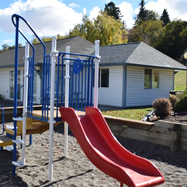 A playground featuring a red slide and climbing structure, set in a sandy area with a background of a house and green grass.