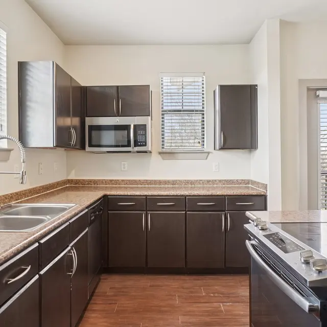 A modern kitchen featuring dark wood cabinets, stainless steel appliances, and a large countertop with a double sink. There are windows with blinds allowing natural light in, and a view outside. The kitchen has a clean and contemporary look.