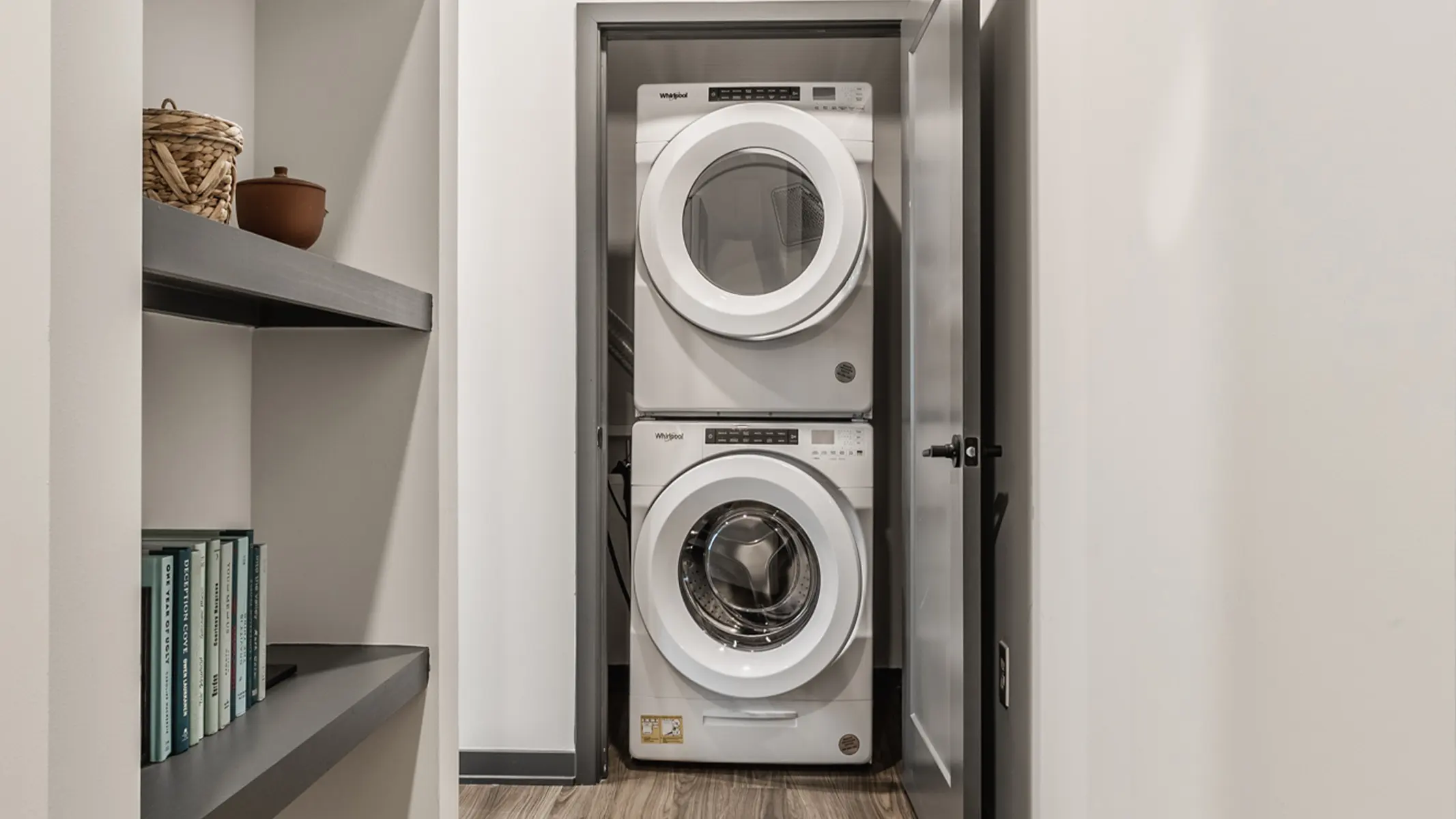 A compact laundry area featuring a stacked washer and dryer unit between a wall and a shelf with books and decorative items.
