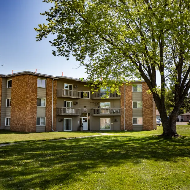 A multi-unit apartment building with brick and siding exterior surrounded by green grass and trees.