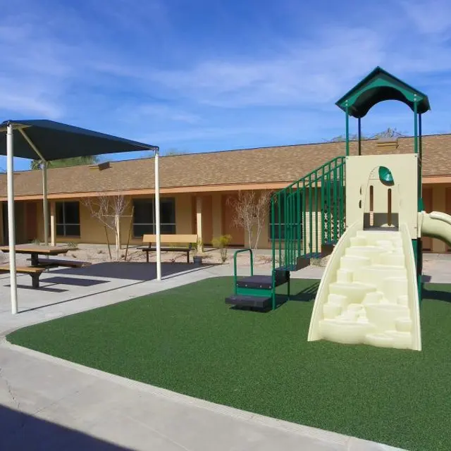 Outdoor Playground Scene An outdoor playground featuring a play structure with a slide, surrounded by a grassy area and benches under a shade structure.