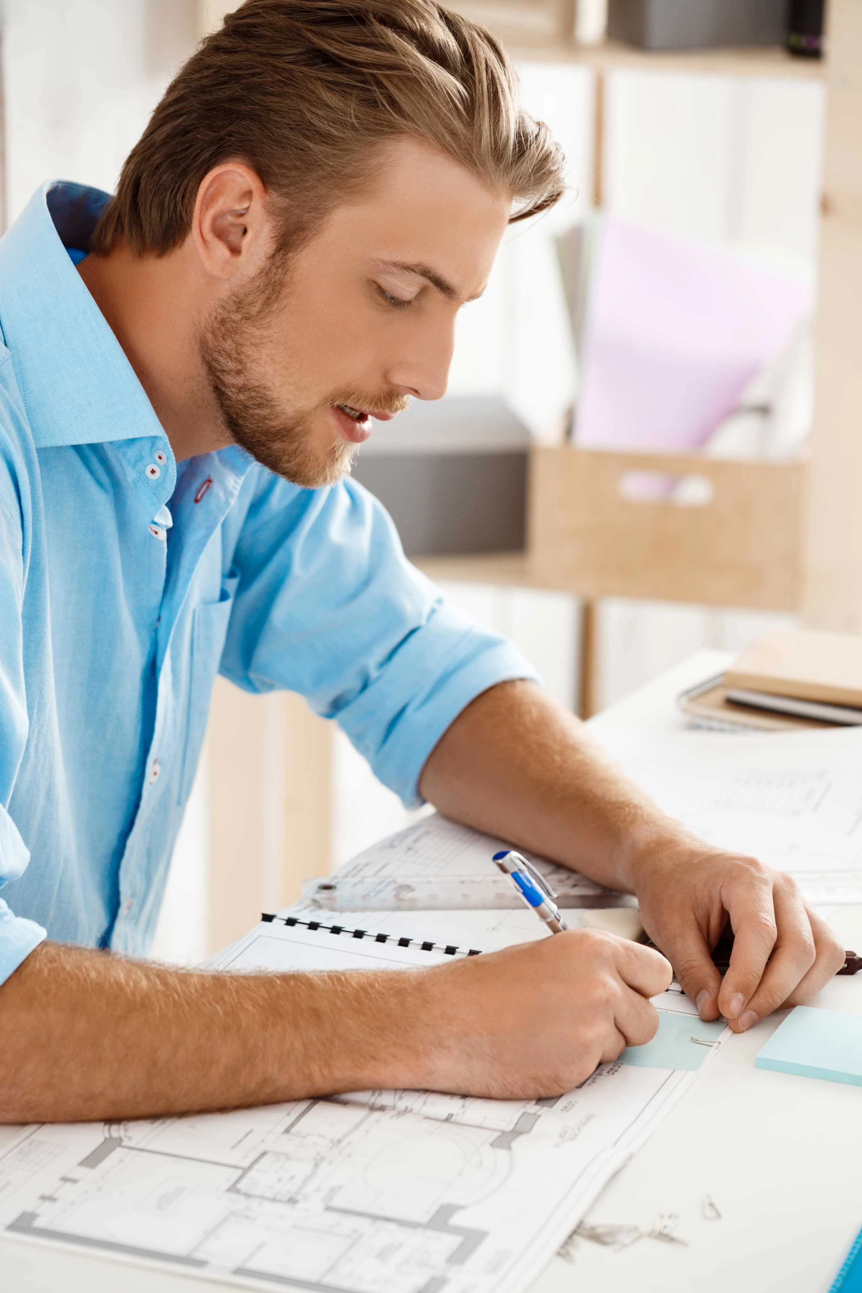 A young man in a blue shirt is focused on drawing and writing notes on a notepad. Architectural plans and tools are spread out on the table before him.