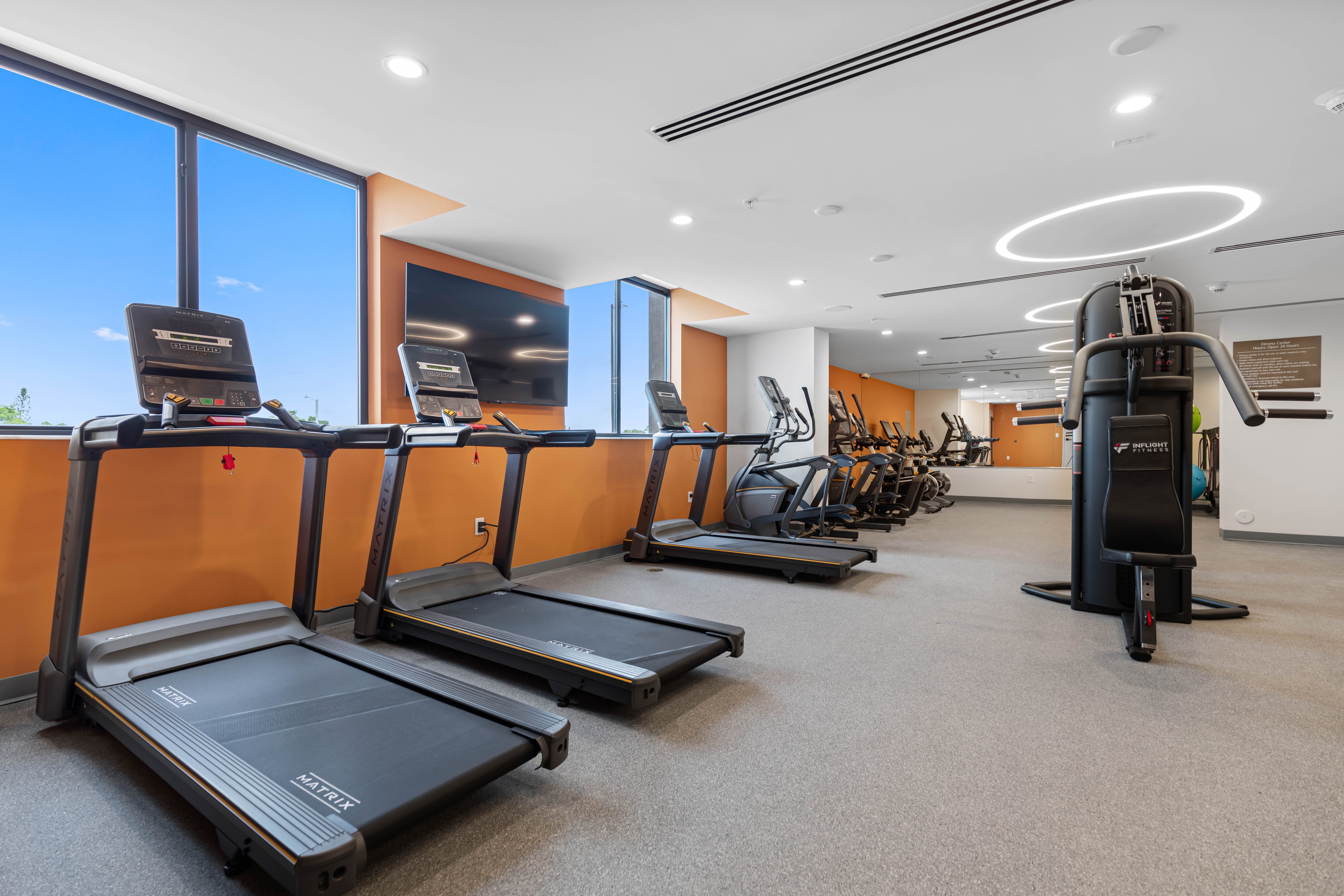Modern Gym Interior A modern gym interior featuring several treadmills, exercise bikes, and a weight machine. Large windows allow natural light to brighten the space, which has a sleek design with orange and neutral tones.