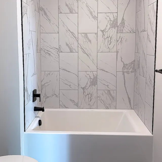 A modern apartment bathroom in richmond va featuring a white bathtub against a marble-patterned tile wall, with black fixtures and a wooden floor.