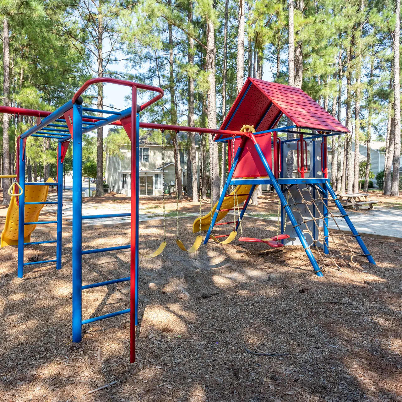 A colorful playground featuring a climbing structure and swings, surrounded by tall trees and a sandy area.