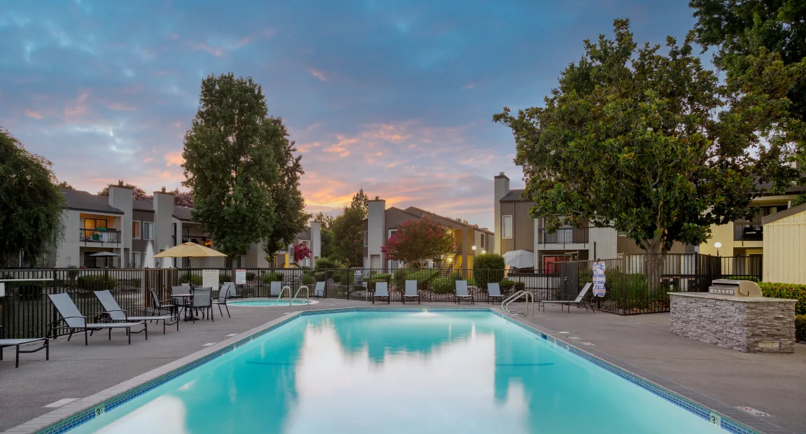 A serene swimming pool surrounded by lounge chairs and lush greenery, with residential buildings in the background and a colorful sky at sunrise.