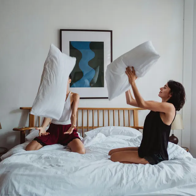 Pillow Fight Between Friends Two people in a cozy bedroom having a pillow fight, with one person holding a pillow above their head and the other person preparing to strike back.