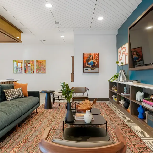 A modern living room with a teal accent wall, a large flat-screen TV, a dark green sofa, and a patterned rug. There are decorative items on shelves and walls adorned with colorful artwork.