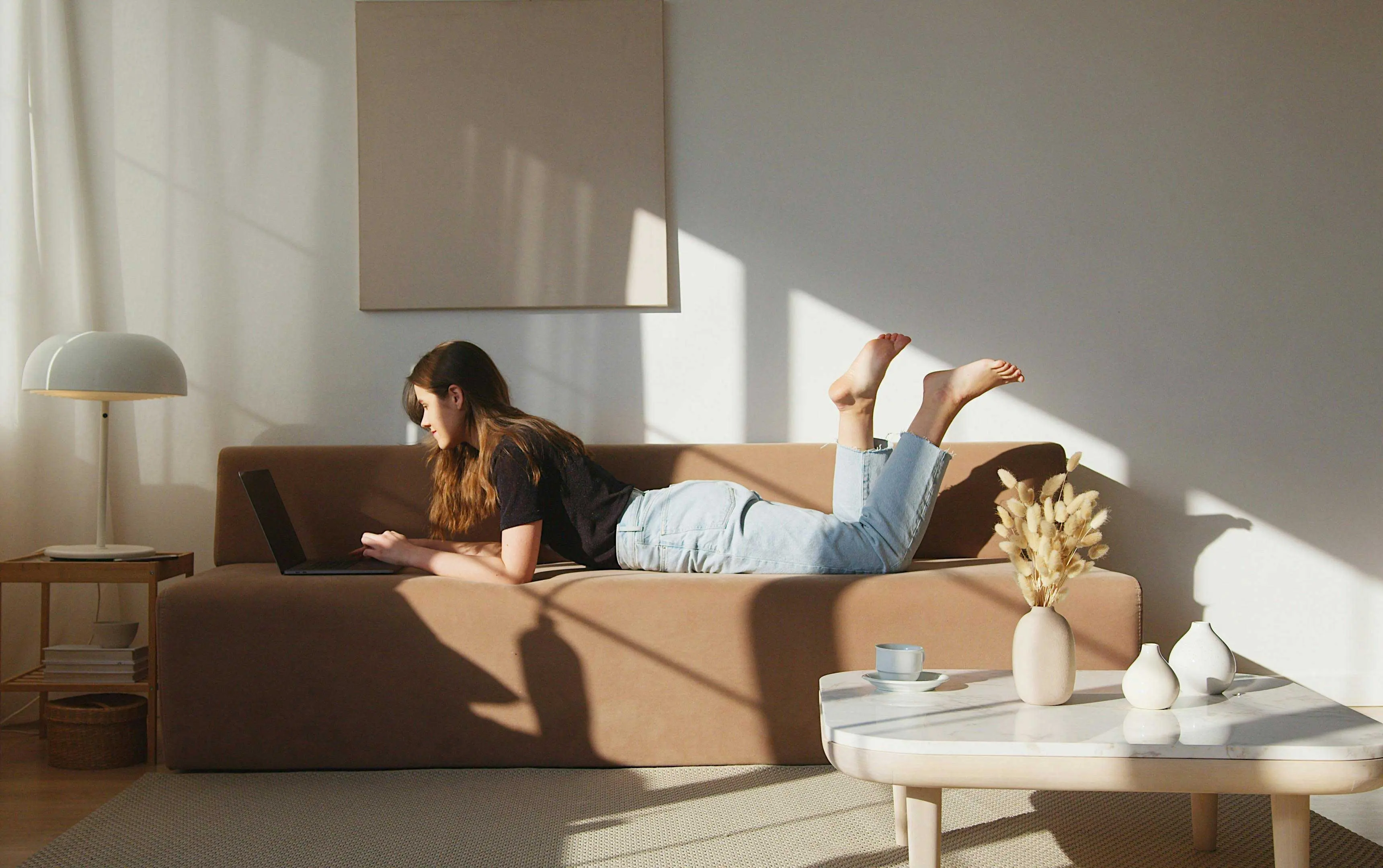 Woman Working on Laptop in Sunlit Living Room A woman lying on a brown sofa, using a laptop. She is propped up on her elbows with her legs raised in the air. The room is filled with natural light, casting soft shadows. Decorative plants and a small table with a cup and vases are visible nearby.