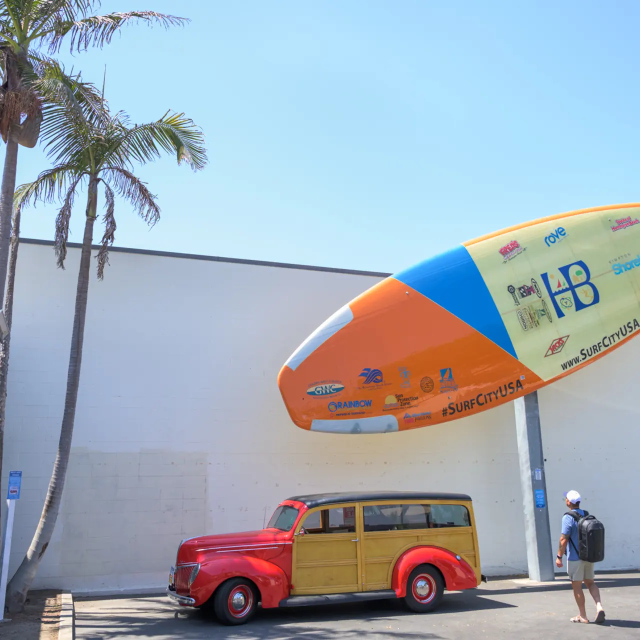 A colorful oversized surfboard mounted above a vintage red car, with a palm tree in the background and a wall decorated with murals.