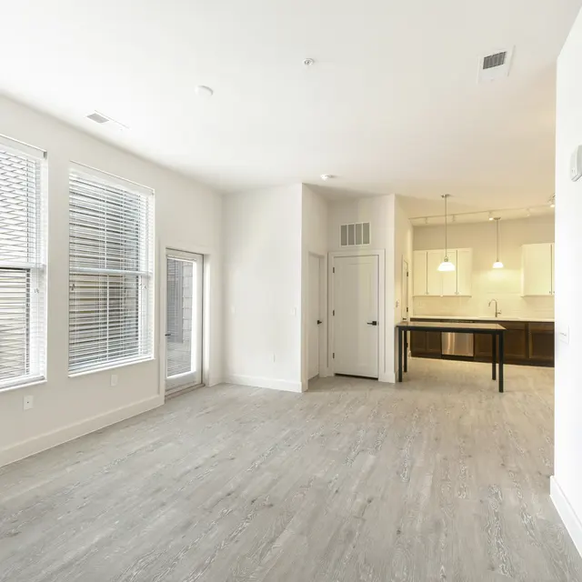 Open Living Space in Modern Apartment A bright, open living space with large windows and light wooden flooring. The room leads to a kitchen area with modern fixtures visible in the background.