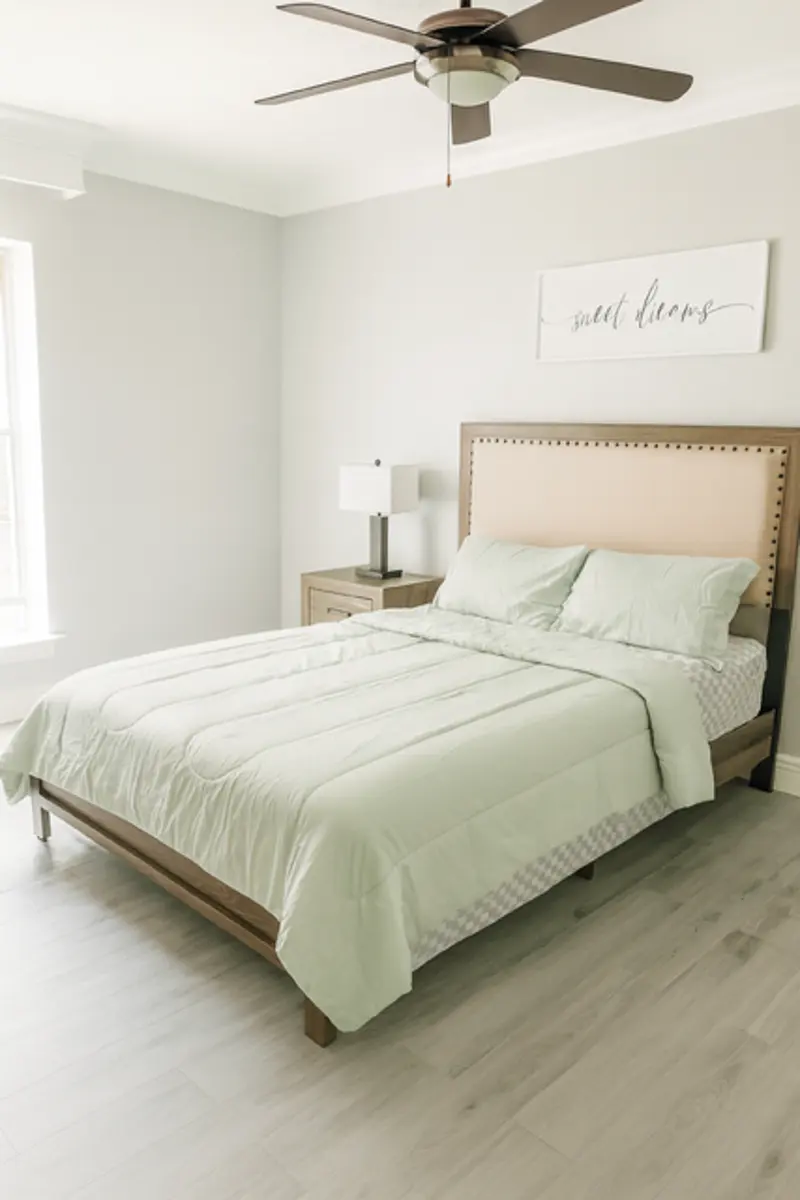 A modern bedroom featuring a light green bedspread on a bed with a beige headboard. There is a table lamp on a nightstand beside the bed, and a wall art piece above the headboard that reads 'sweet dreams'.