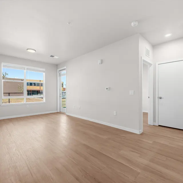 A spacious, empty living room with light-colored walls and hardwood flooring. Large windows allow natural light to fill the room. Multiple doorways lead to adjoining rooms, and there is a small kitchenette area visible.