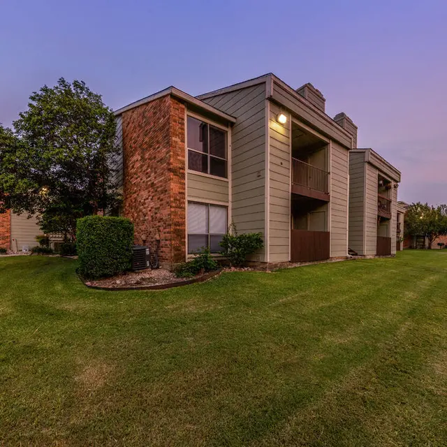 Two-story apartment buildings with a grassy area, landscaped with bushes and trees under a twilight sky.