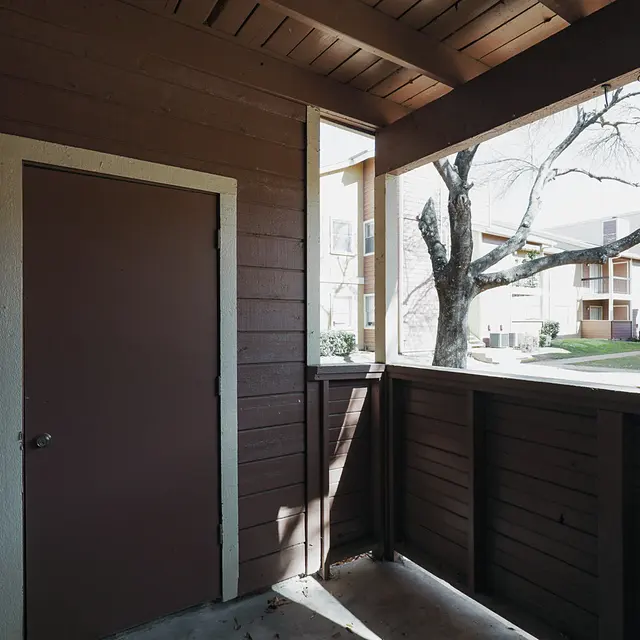 A porch with brown walls and a closed door, featuring a tree and apartment buildings visible in the background.