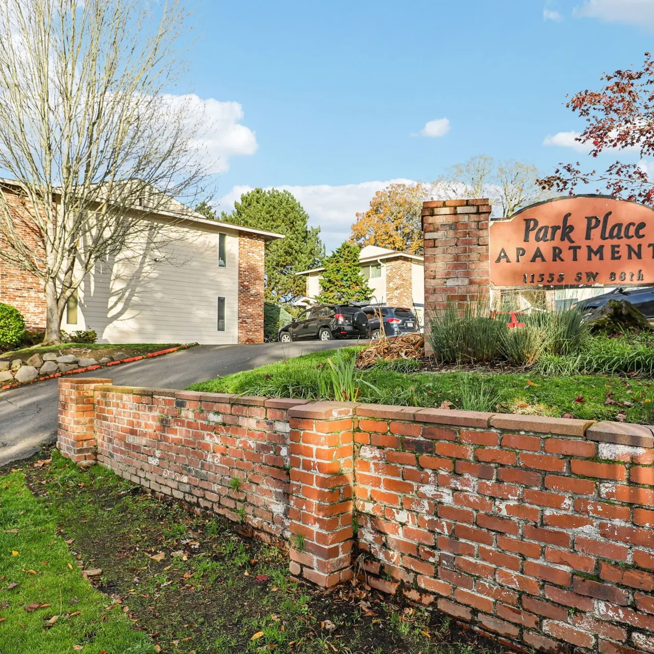 A brick entrance to Park Place Apartments featuring a large sign that reads 'Park Place Apartments'. The scene includes a well-kept lawn with trees and a driveway leading into the apartment complex.