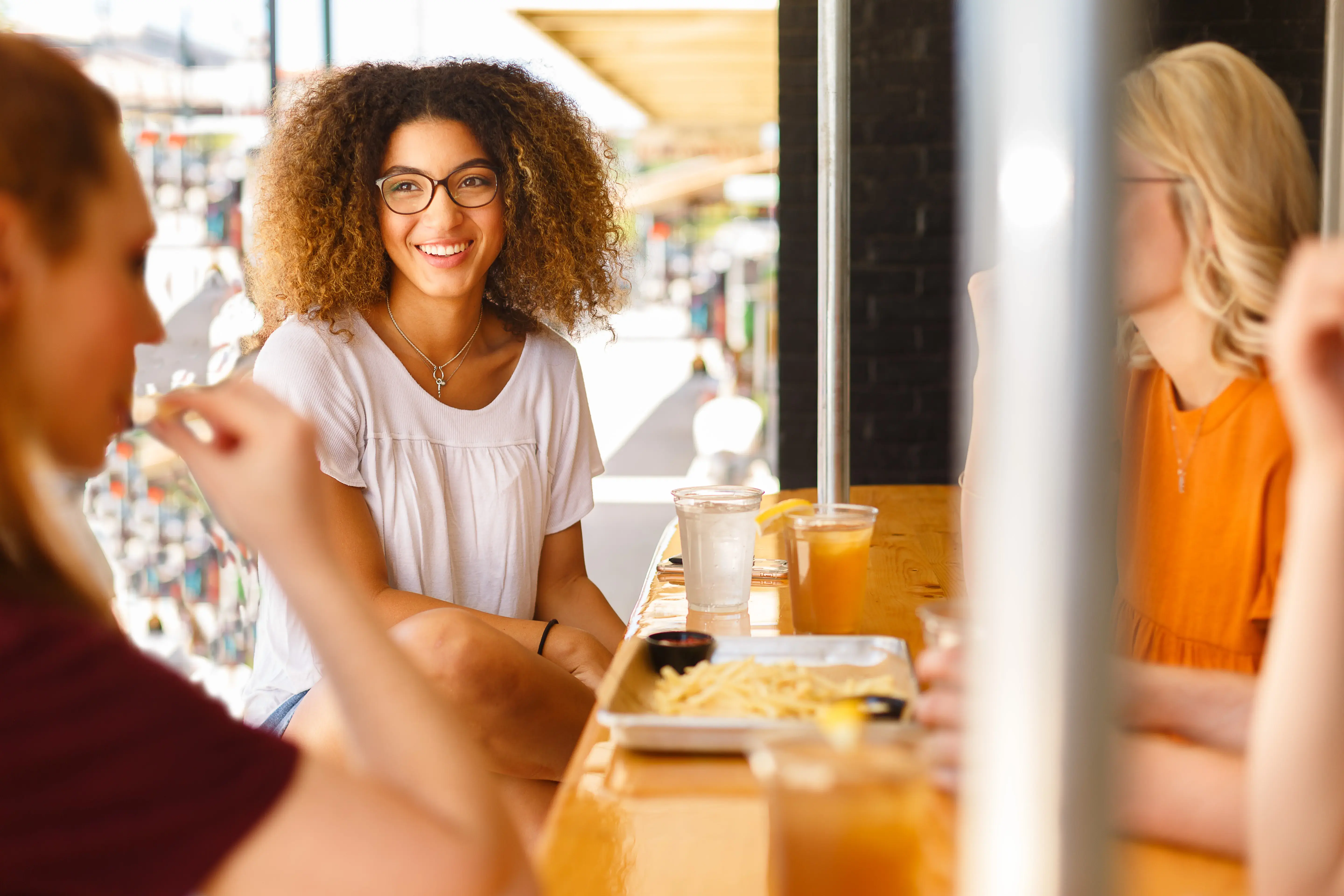 Friends Enjoying Lunch A young woman with curly hair and glasses smiles at the camera while sitting at a wooden table with friends. The table has food and drinks, and they are in a vibrant, casual dining setting.