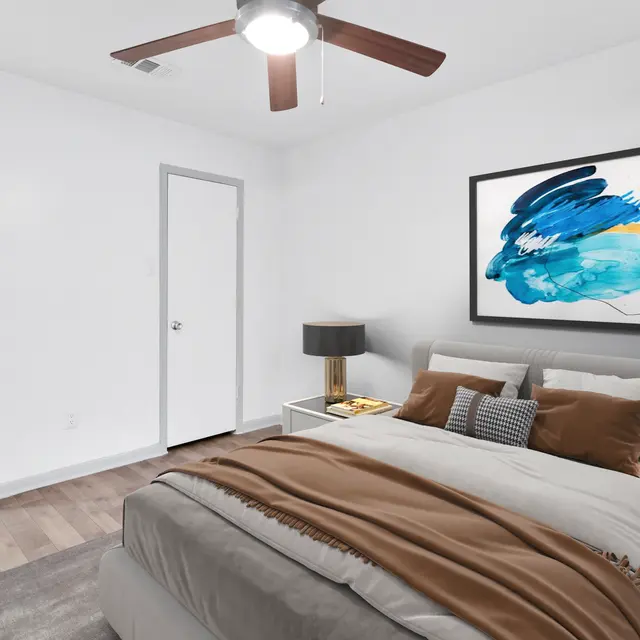 A modern bedroom featuring a king-sized bed with a soft gray blanket and patterned cushions. The room has white walls, wooden flooring, and a ceiling fan. There is a piece of abstract art hanging above the bed, and two bedside tables with black lamps. A door leads to another room.