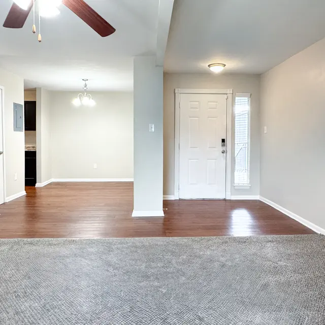 A spacious living area with hardwood floors, a light-colored wall, and an entrance door leading outside. There are light fixtures and a ceiling fan visible. To the left, there's a view of another room in the background.