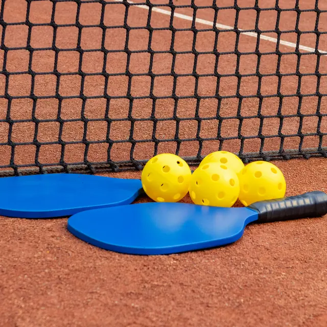 Two blue paddles and four yellow pickleballs resting on a red clay court next to a net.