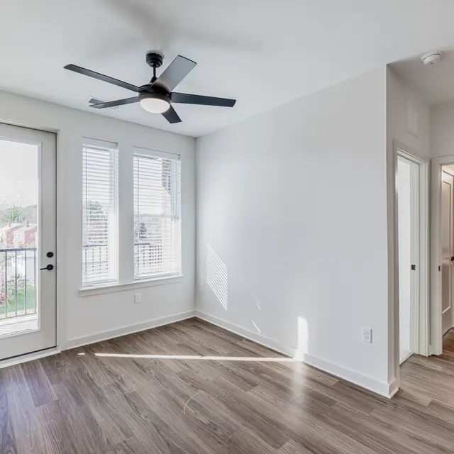 A bright and airy room in a modern apartment featuring a ceiling fan, wooden flooring, and large windows with a door opening to a balcony. The room is connected to a bathroom area through an open door.