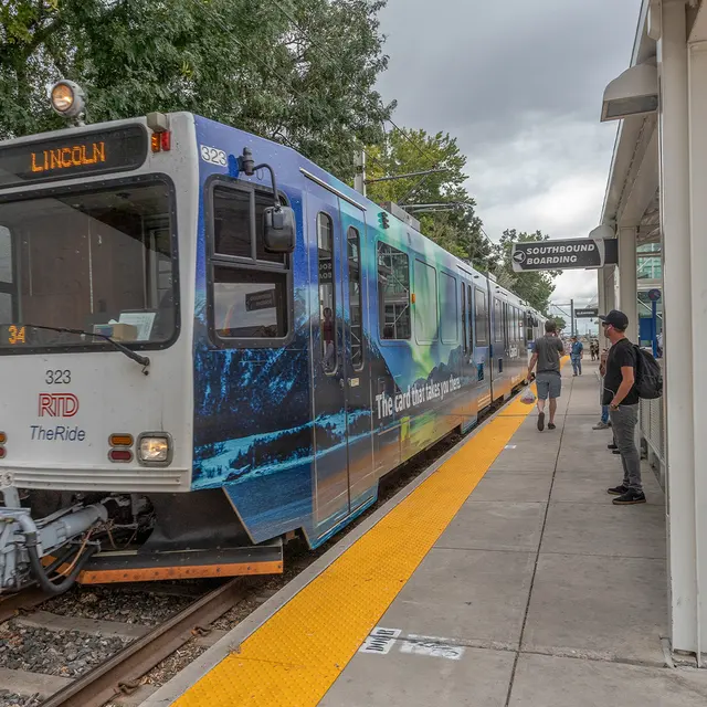A light rail train at a station with passengers waiting. The train features a colorful blue and green design, and the word 'LINCOLN' displayed on the front. The platform is partially covered with a roof, and trees are visible in the background.