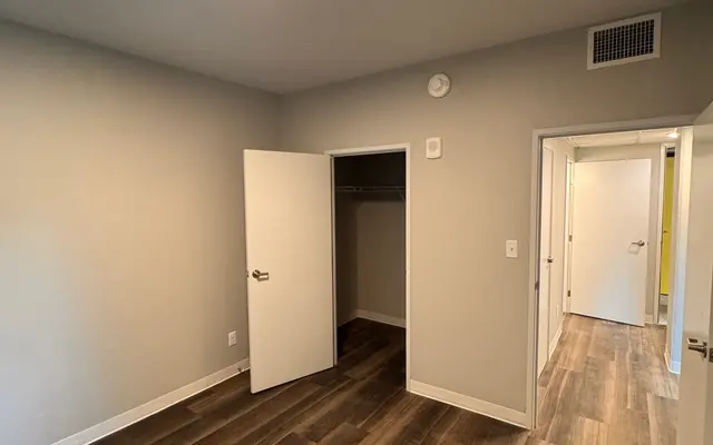 Empty Room Interior An empty room with light gray walls and hardwood flooring. There are two open doors leading to closets and another door visible in the background. A ceiling light is illuminating the space.