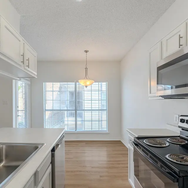 A modern kitchen featuring a stainless steel stove, microwave, and a double sink with a window above it. Natural light floods the space through two large windows. The flooring is a light wood finish, and there is a hanging light fixture over the sink area.