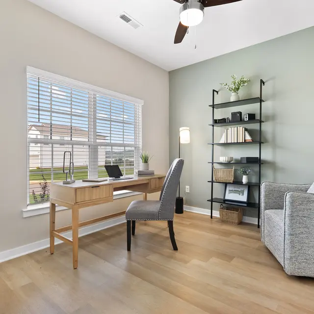 A cozy home office setup featuring a wooden desk, a grey chair, a bookshelf, and a window with white blinds letting in natural light.