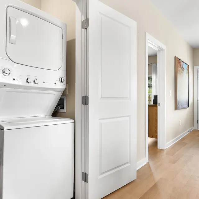 A compact laundry area featuring a stackable washer and dryer, located in a hallway. The walls are light-colored, and there are doors leading to other rooms.