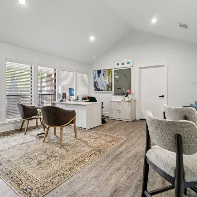 A modern office interior featuring a wooden floor, a large rug, two brown chairs, and a white desk. Sunlight filters through multiple large windows lined with blinds. A colorful piece of art hangs on the wall, adding a creative touch to the space.