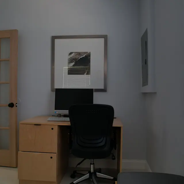 Interior view of a simple office space featuring a wooden desk with a computer, a black chair, and a glass door in the background.