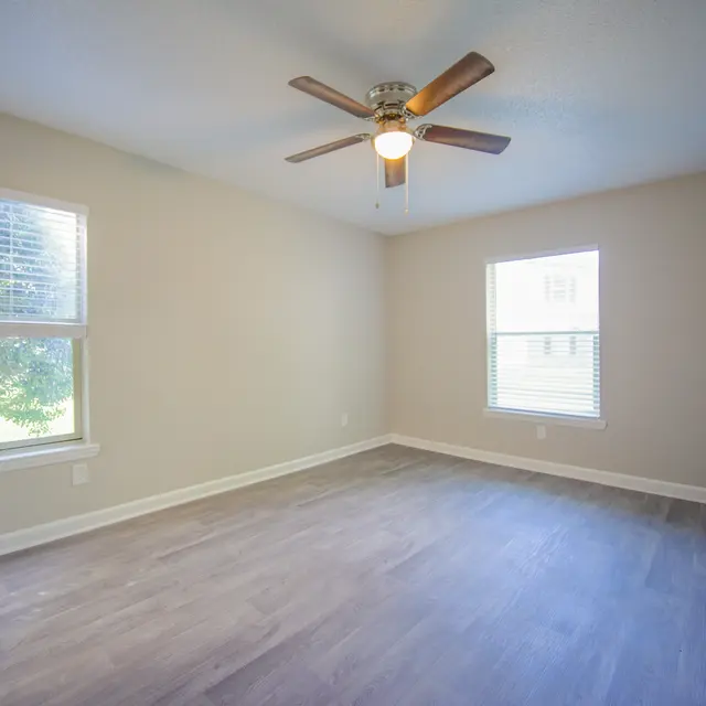 An empty bedroom featuring a ceiling fan, with two windows allowing natural light, and a light wooden floor.